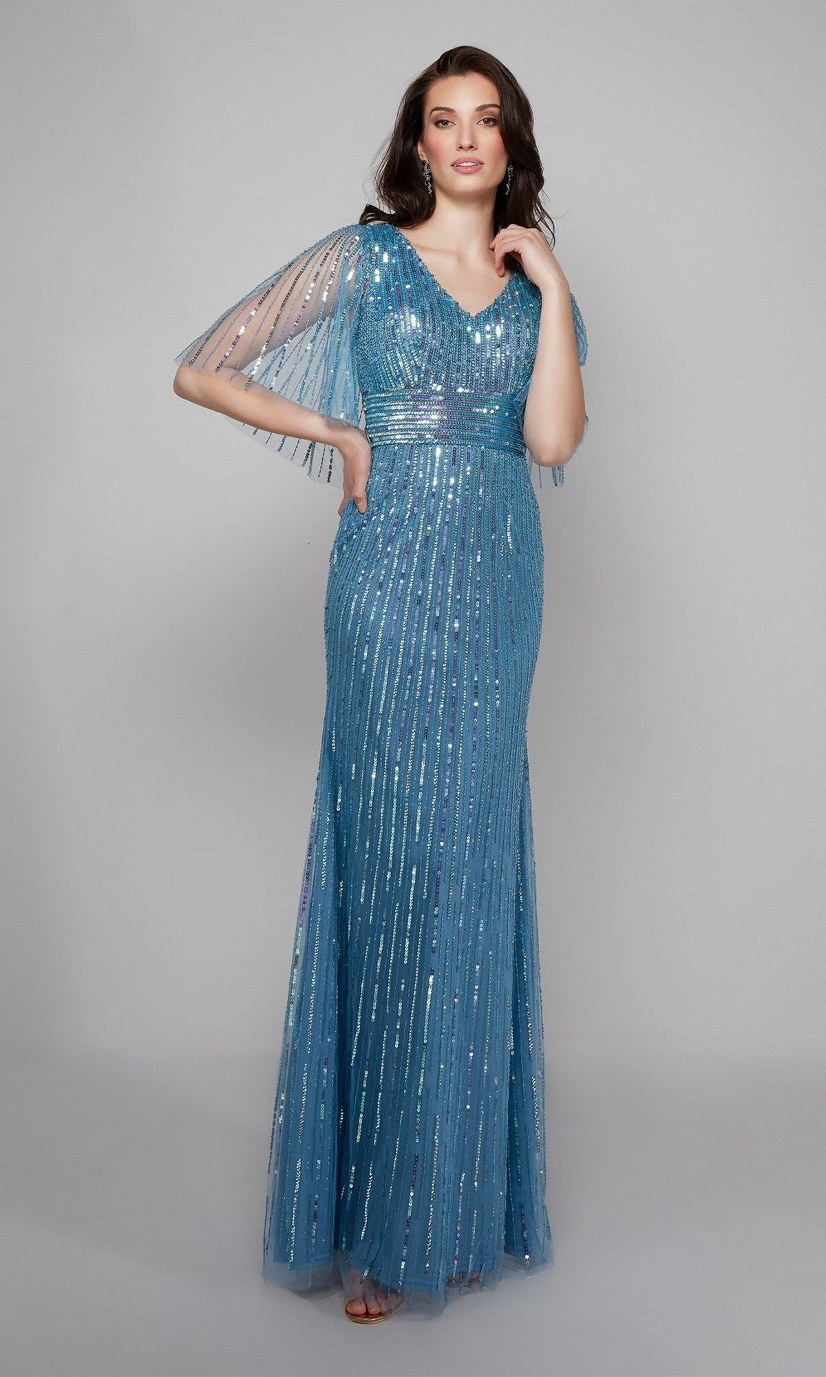 A woman poses in the Alyce 27599, a long formal beaded sequin dress in blue with vertical detailing, sheer short sleeves, and a V-neckline—an elegant mother-of-the-bride gown set against a plain background.
